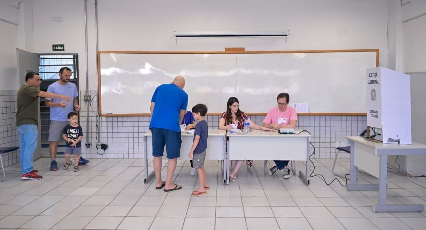 Foto mostra dois eleitores e três mesários em uma seção eleitoral