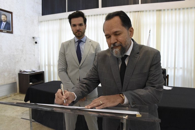 Desembargador Ramom Tácio assina um documento. O deputado Tadeu Leite está ao fundo, observando.