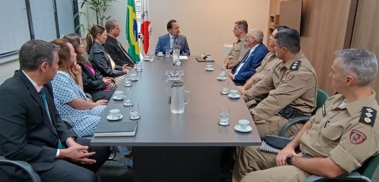 Representantes do TRE-MG e da Polícia Militar sentados ao redor de uma mesa retangular. Ao fundo...