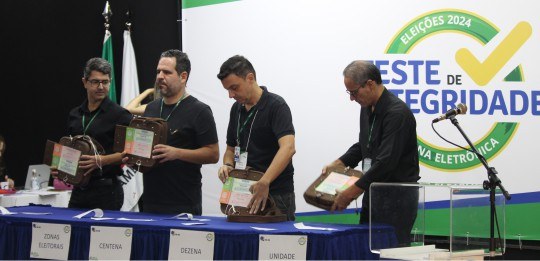 Foto mostra quatro homens de pé junto a uma mesa. Os quatro seguram urnas de lona.