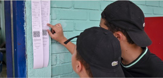 Foto em close de dois rapazes de boné preto observando boletins de urna colados na parede.