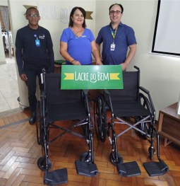 Uma mulher e dois homens estão de pé junto a duas cadeiras de rodas. Eles seguram um cartaz verd...