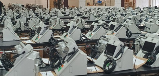 Foto mostra dezenas de urnas eletrônicas sobre bancadas.