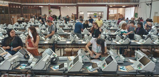 Foto mostra dezenas de urnas eletrônicas sobre bancadas e algumas pessoas de pé perto dos equipa...