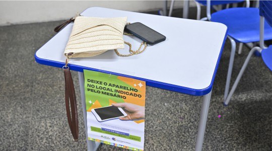 Foto mostra uma bolsa bege e um celular sobre uma mesa. Na frente da mesa, há um cartaz com a fr...