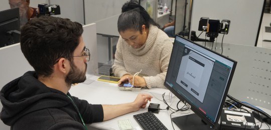 Rapaz observa uma mulher assinando em equipamento digital, assinatura aparece na tela do computa...