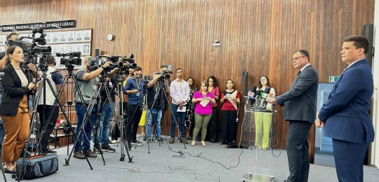 No lado esquerdo da foto, diversos jornalistas de pé e algumas câmeras sobre tripés. Do lado dir...