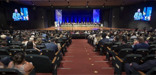 Foto mostra um grande auditório com a plateia em primeiro plano. Ao fundo, o desembargador Ramom...