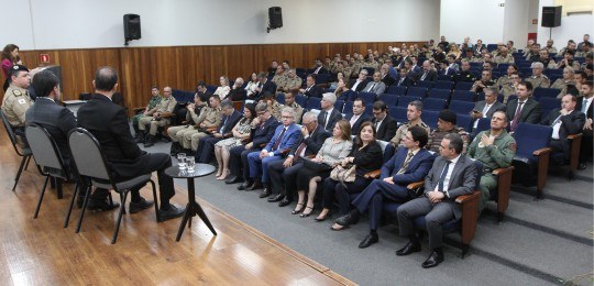 Em primeiro plano, diversas pessoas sentadas na plateia de um auditório, de costas para a câmera...