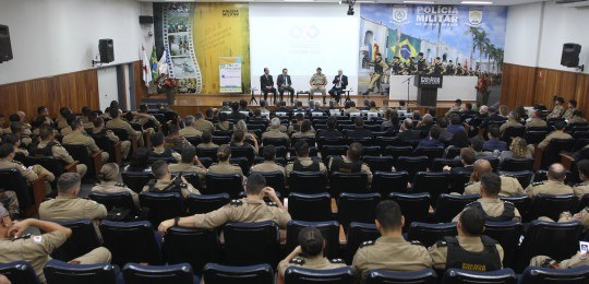 Em primeiro plano, diversas pessoas sentadas na plateia de um auditório, de costas para a câmera...