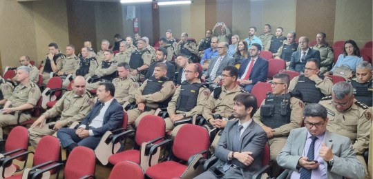 Em primeiro plano, diversas pessoas sentadas na plateia de um auditório, de costas para a câmera...