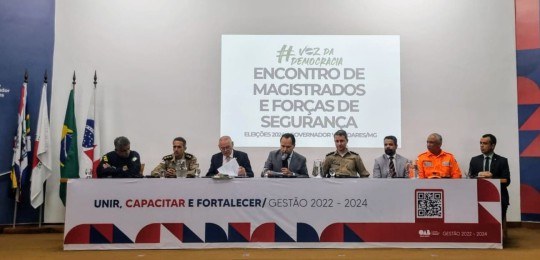 Em primeiro plano, diversas pessoas sentadas na plateia de um auditório, de costas para a câmera...