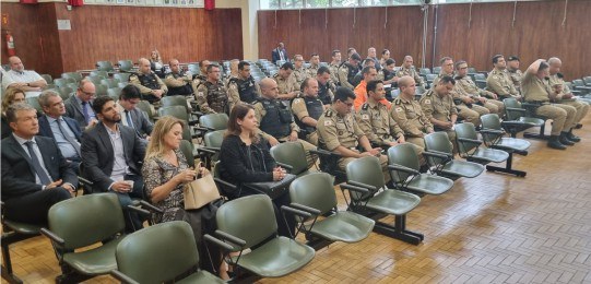 Em primeiro plano, diversas pessoas sentadas na plateia de um auditório, de costas para a câmera...