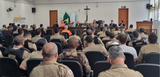 Em primeiro plano, diversas pessoas sentadas na plateia de um auditório, de costas para a câmera...