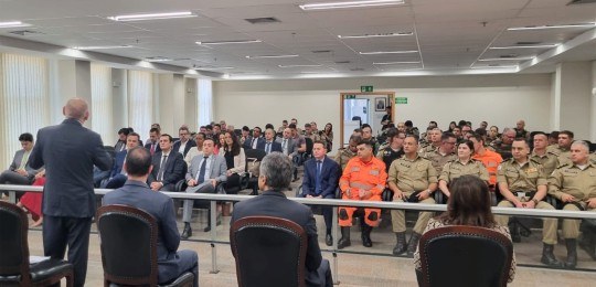 Em primeiro plano, diversas pessoas sentadas na plateia de um auditório, de costas para a câmera...