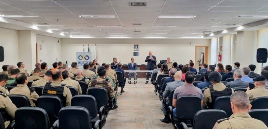 Em primeiro plano, diversas pessoas sentadas na plateia de um auditório, de costas para a câmera...