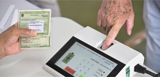 Foto mostra um homem colocando o dedo indicador da mão direita no leitor biométrico do terminal ...