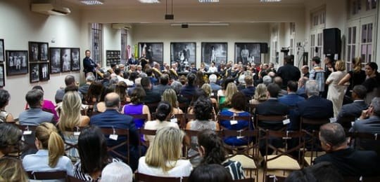 Diversas pessoas sentadas em uma plateia, de costas para a câmera. Ao fundo, autoridades sentada...