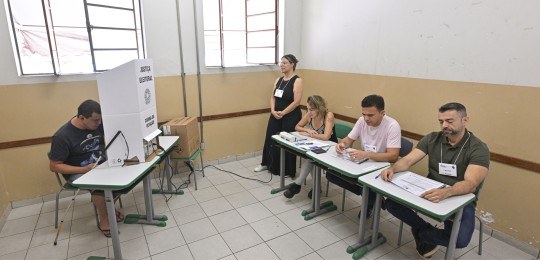 Foto mostra um eleitor cego, com bengala, na cabine de votação. No canto direito da foto, aparec...