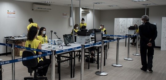 Pessoas usando coletes amarelos estão sentadas junto a urnas eletrônicas e monitores de TV. Um h...