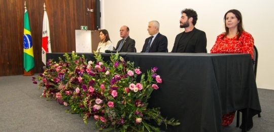 Foto de uma mesa onde estão sentadas autoridades, tendo a frente um arranjo de flores