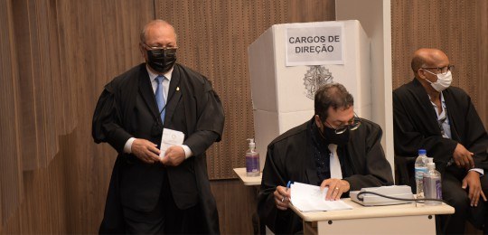 Foto de cabine de votação atrás de um homem sentado perto de uma mesa. Há dois homens de pé, um ...