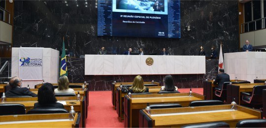 Foto mostra algumas quatro homens em uma mesa e, em primeiro plano, algumas pessoas sentadas no ...