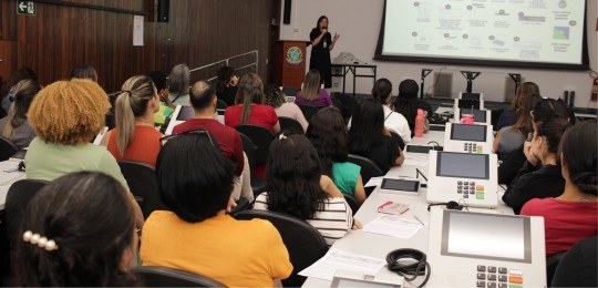 Foto de um auditório. Em primeiro plano, pessoas sentadas, de costas para a câmera. Ao fundo, um...