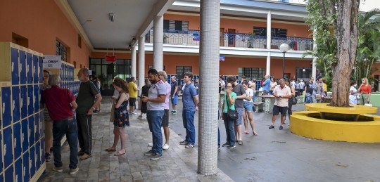 Foto mostra eleitoras e eleitores em fila na porta de uma seção eleitoral e andando pelo pátio d...