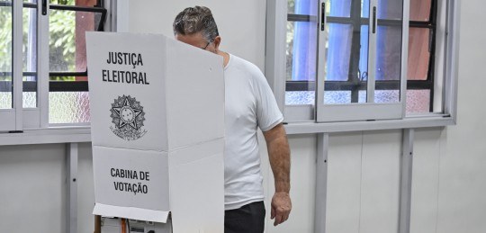 Foto de um homem atrás de uma cabine de votação com o brasão da Justiça Eleitoral.